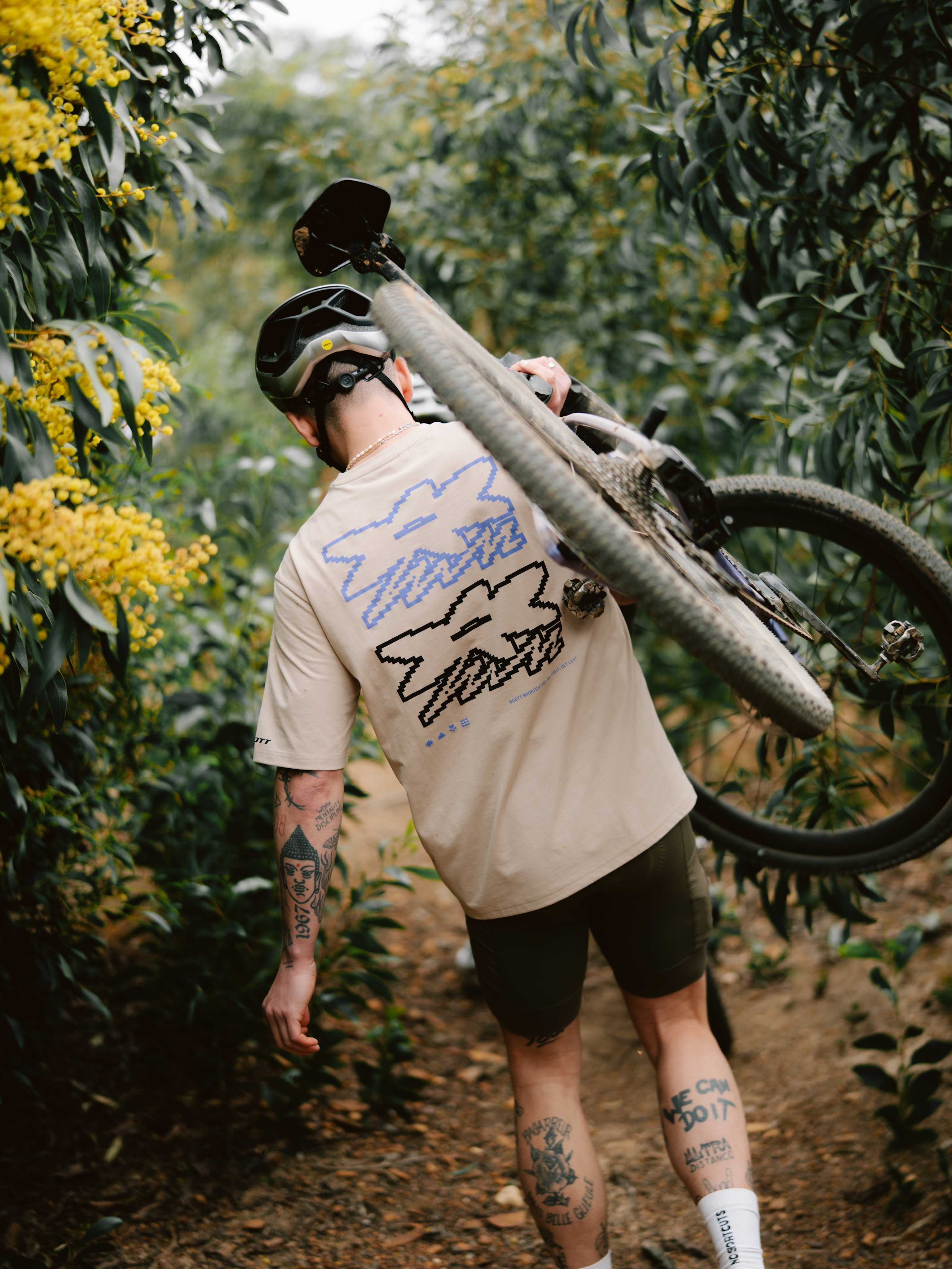 A man in a tan t-shirt and helmet carries a mountain bike on his shoulder through a wooded trail with yellow flowers on the left and dense green foliage in the background.