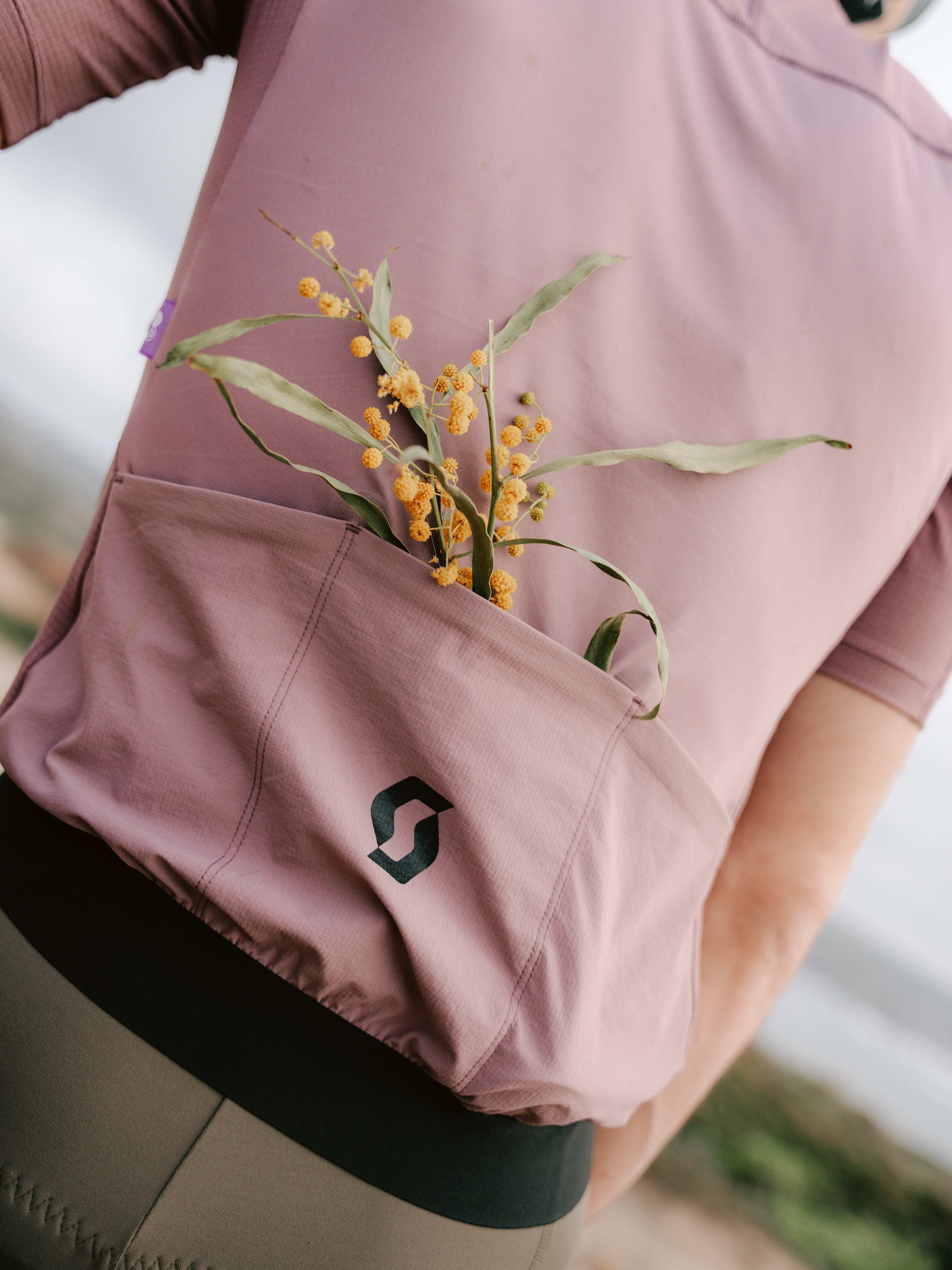 A sprig of yellow wildflowers peeks from the back pocket of a cyclist's mauve jersey, with a blurred, outdoor landscape in the background.