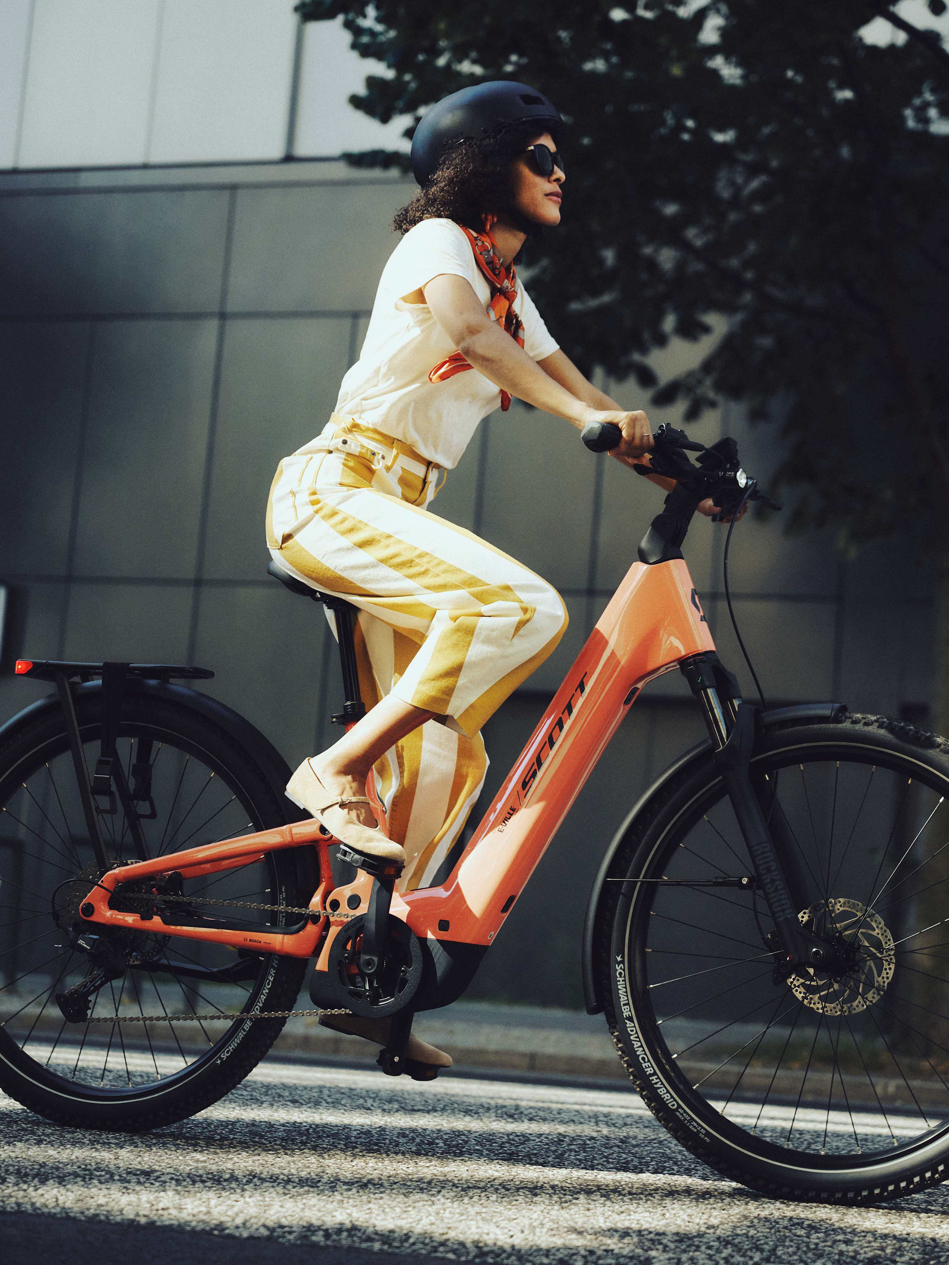 A woman in striped pants rides an orange electric bicycle on a street with a modern building and trees in the background.