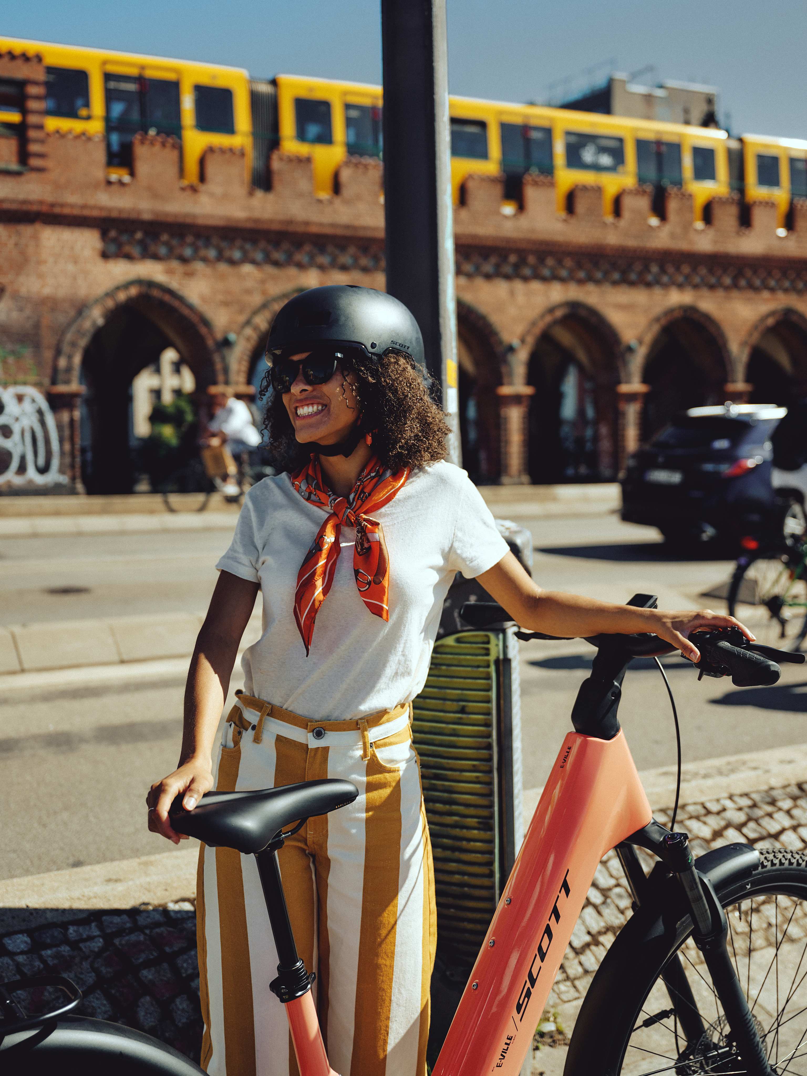 A smiling woman in a white shirt and striped pants stands with her bicycle in the foreground, while a yellow train passes over an arched brick bridge in the background.