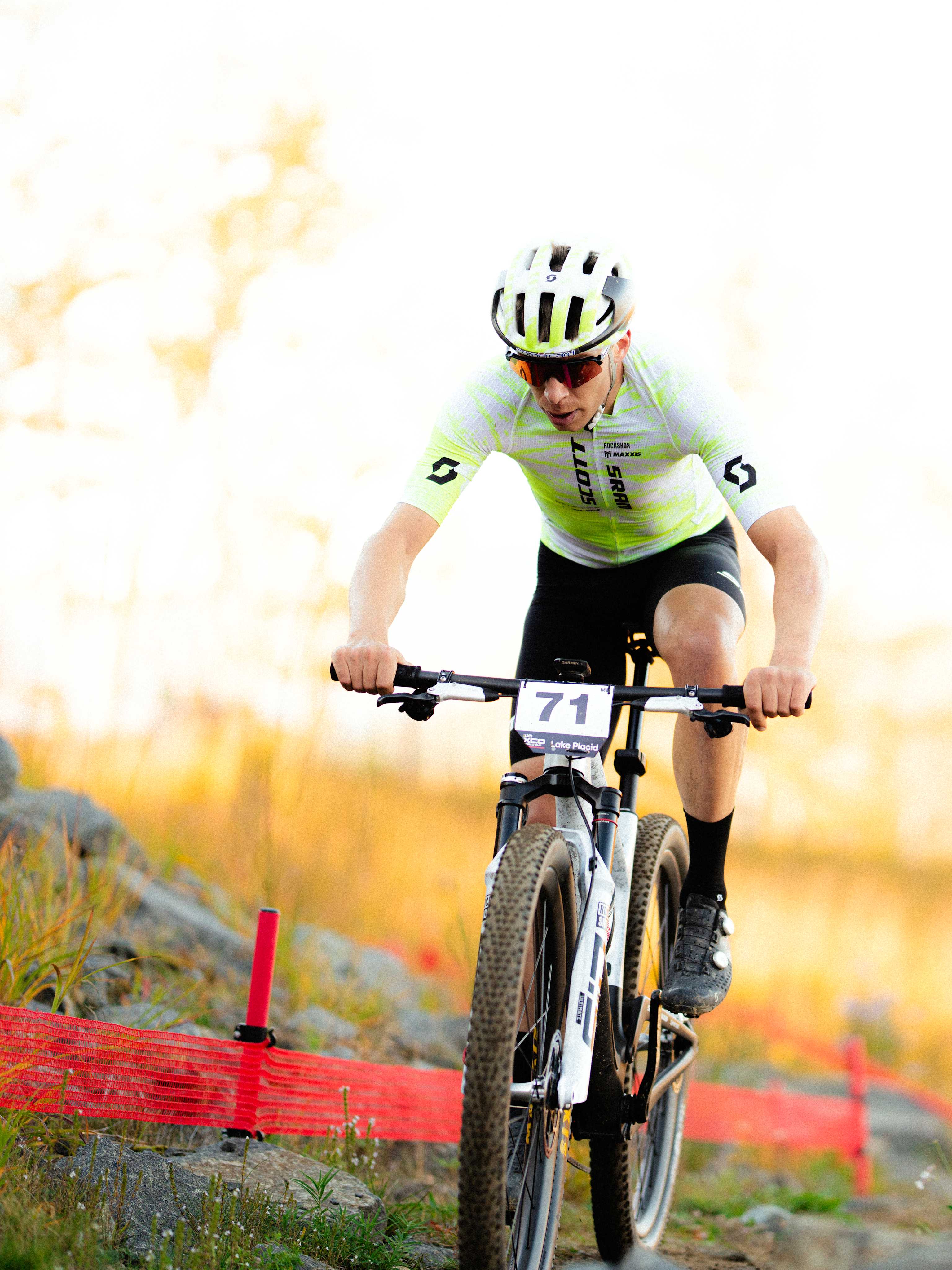 A cyclist in a neon yellow jersey and white helmet rides a mountain bike along a rocky trail bordered by red fencing against a blurred autumn forest background.