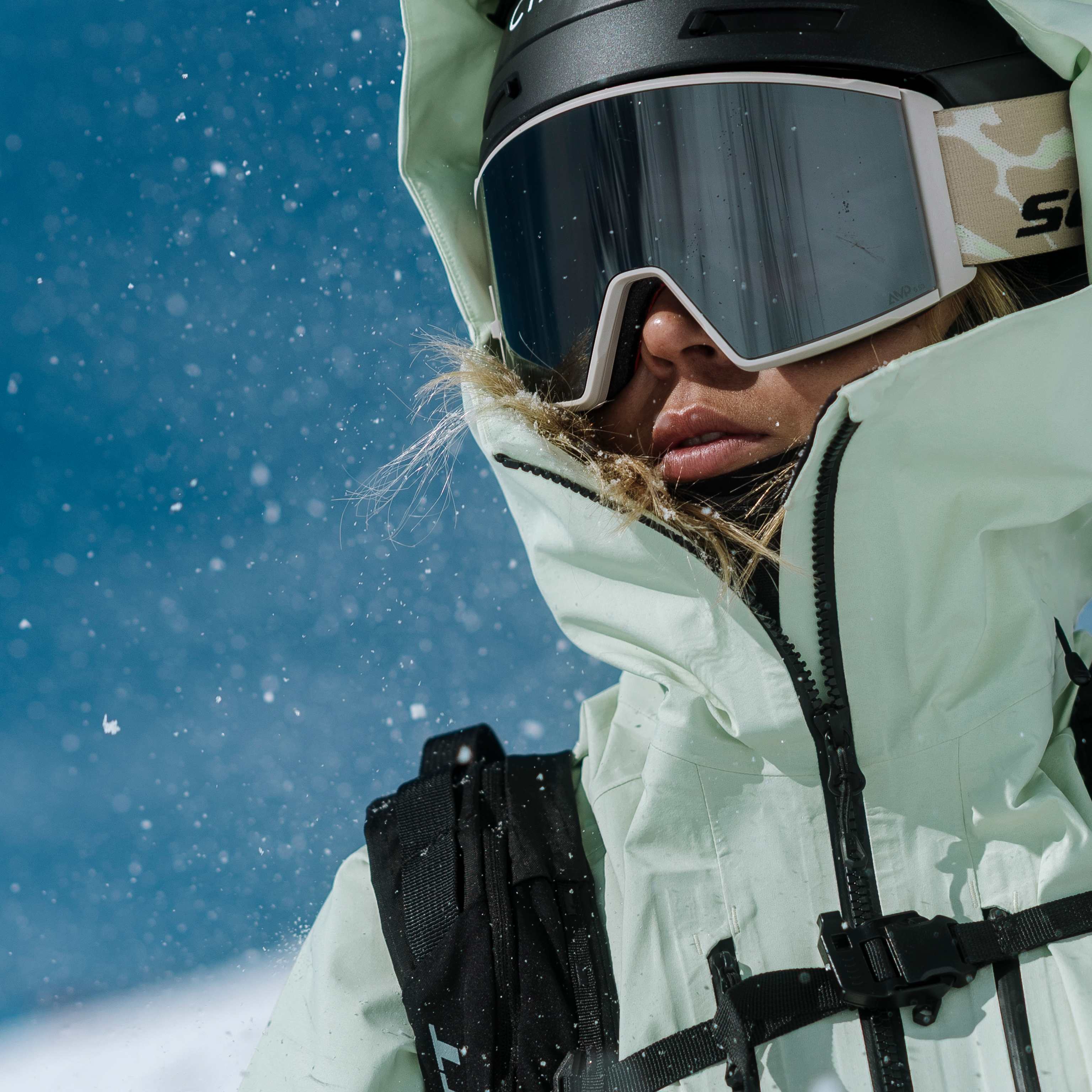 A skier in a light green jacket and goggles is in the foreground, with a snowy mountain slope in the blurred background.