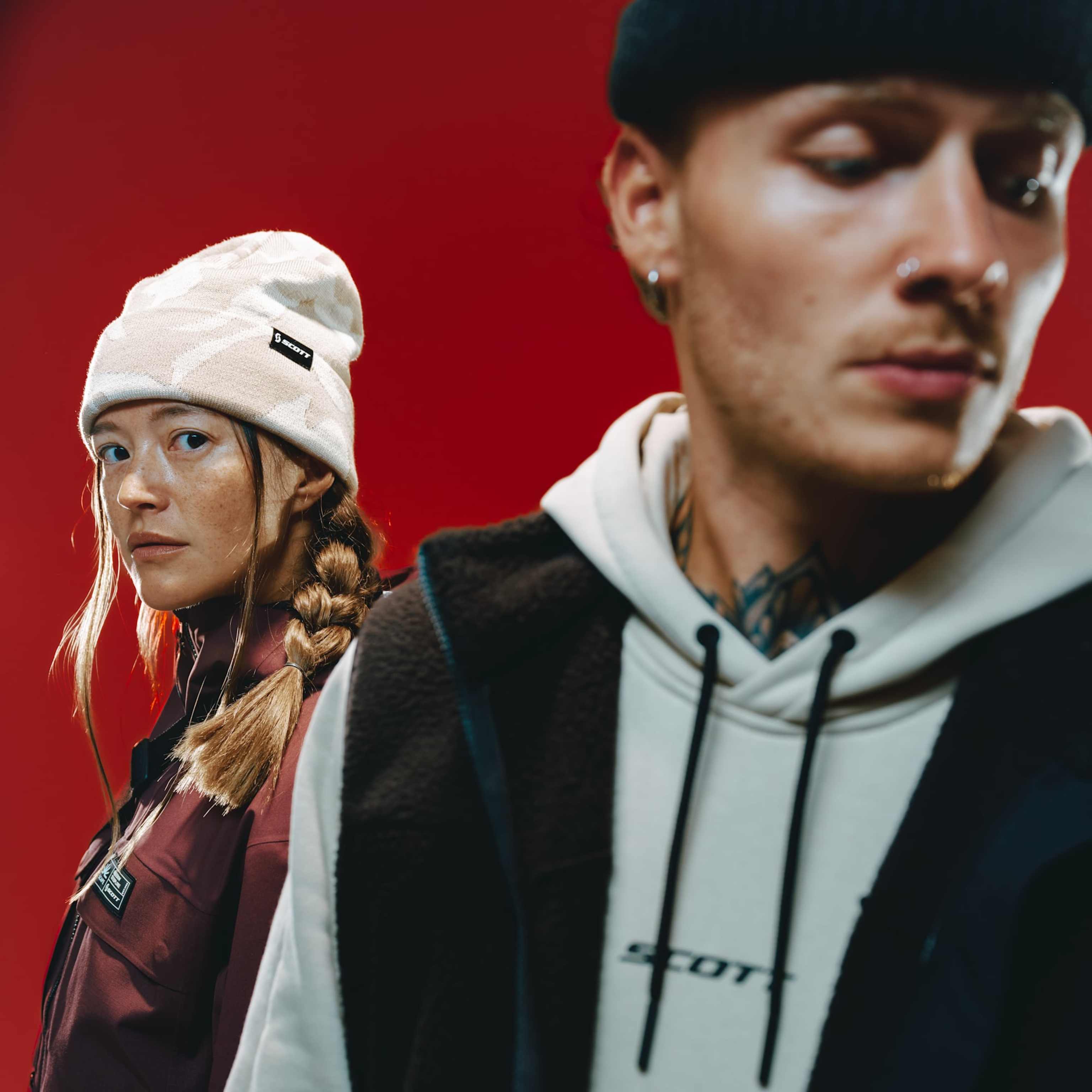 A woman in a white beanie and maroon jacket stands in the background while a man in a black beanie and white hoodie is blurred in the foreground against a red backdrop.