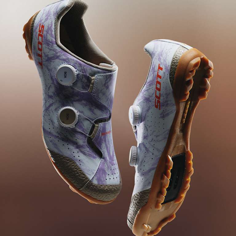 A pair of purple and white marbled cycling shoes with orange "SCOTT" logos are suspended against a softly blurred brown background.