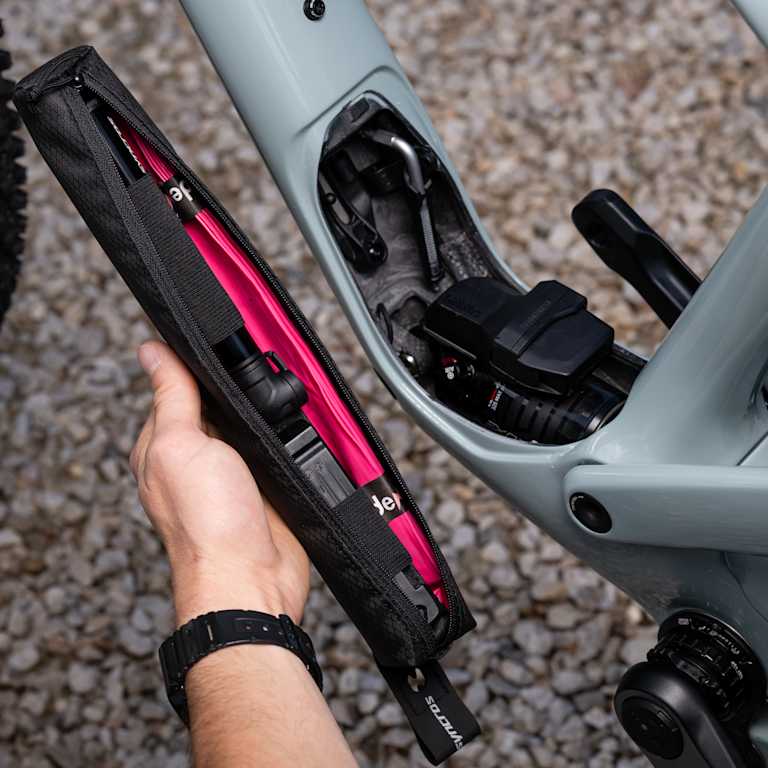 A hand holds a black and pink tool bag near the open compartment of a light gray bicycle frame, with a portion of the rear wheel visible in the blurred background.