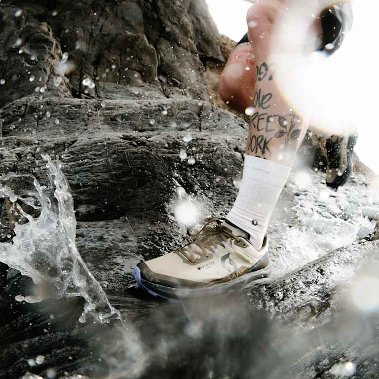 A person's tattooed leg and foot in a tan shoe splash through rocky, sunlit water.