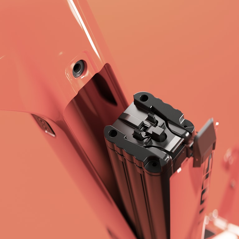 Close-up of a coral-red bicycle frame with black suspension forks and a detailed folding hinge mechanism, conveying precision and modern design.
