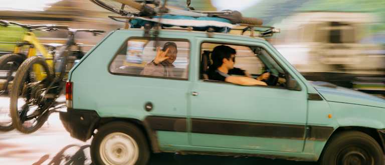 A light blue car with bicycles on its roof and back drives past a blurred background of trees and buildings.
