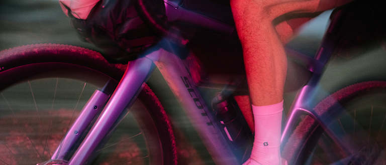 A cyclist in pink socks and white shoes pedals a purple bike with a blurred background of a dusty path.