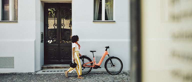 A woman in striped pants walks past a peach-colored bicycle parked in front of a white building with a dark wooden door.