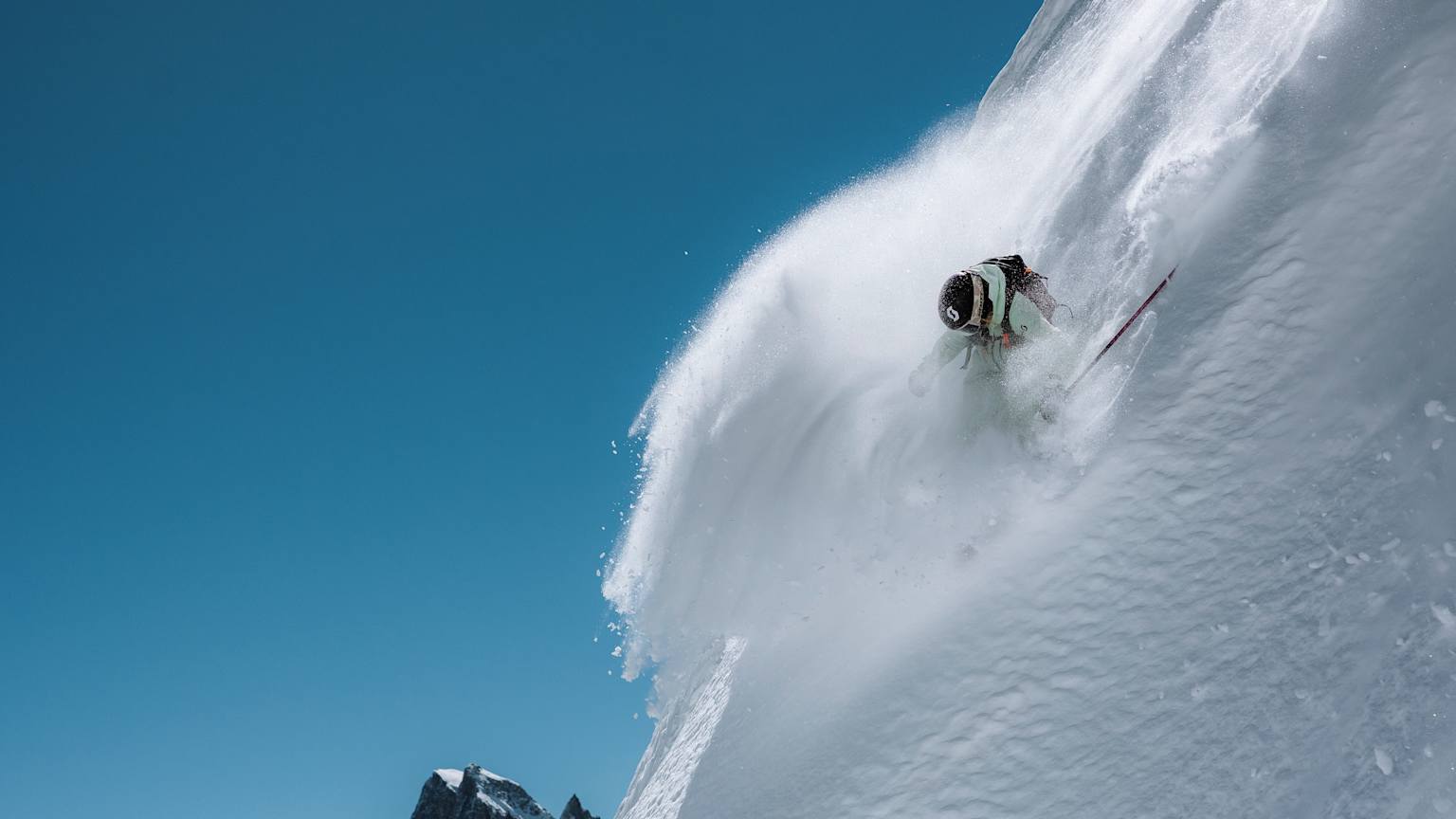 A skier carves down a steep, snow-covered mountain face, kicking up a plume of powder against a clear blue sky and jagged peaks in the distance.