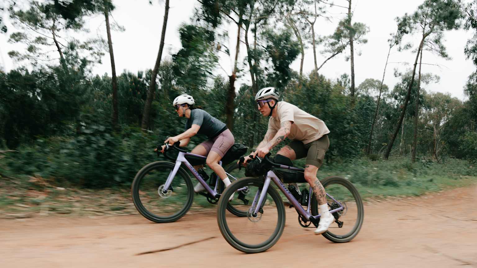 Two cyclists ride gravel bikes on a dirt path with trees in the background.
