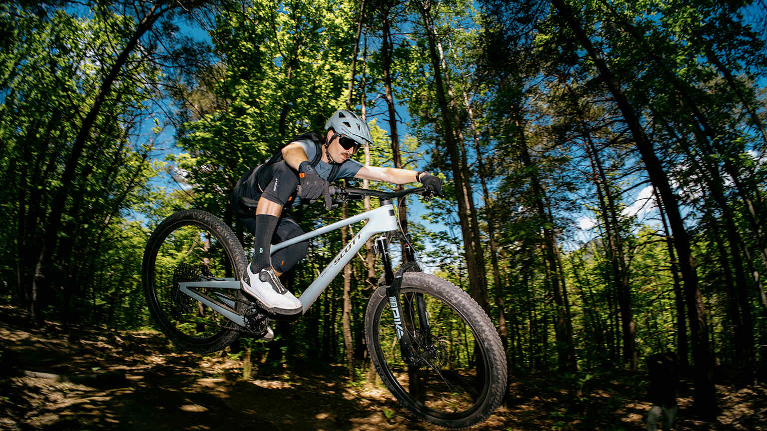 A mountain biker in the foreground rides down a dirt trail surrounded by a lush green forest in the background.