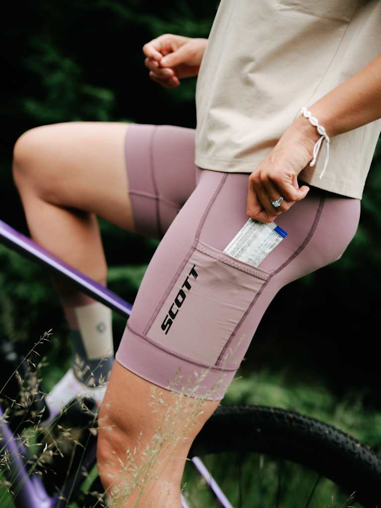 A person in mauve cycling shorts with a gel pack in the pocket sits on a purple bicycle with a blurred green forest in the background.