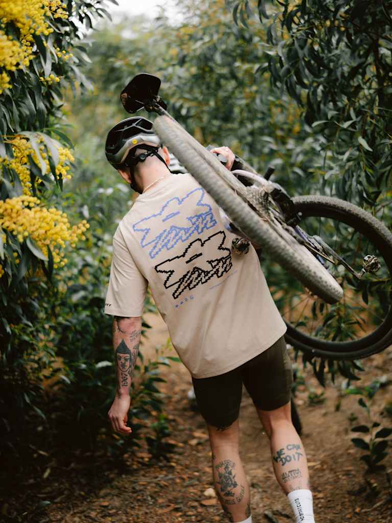 A man in a tan t-shirt and helmet carries a mountain bike on his shoulder through a wooded trail with yellow flowers on the left and dense green foliage in the background.