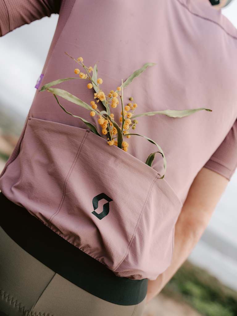 A sprig of yellow wildflowers peeks from the back pocket of a cyclist's mauve jersey, with a blurred, outdoor landscape in the background.