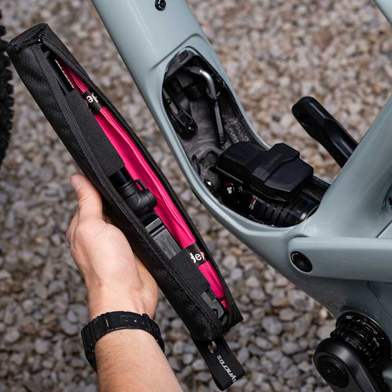 A hand holds a black and pink tool bag near the open compartment of a light gray bicycle frame, with a portion of the rear wheel visible in the blurred background.