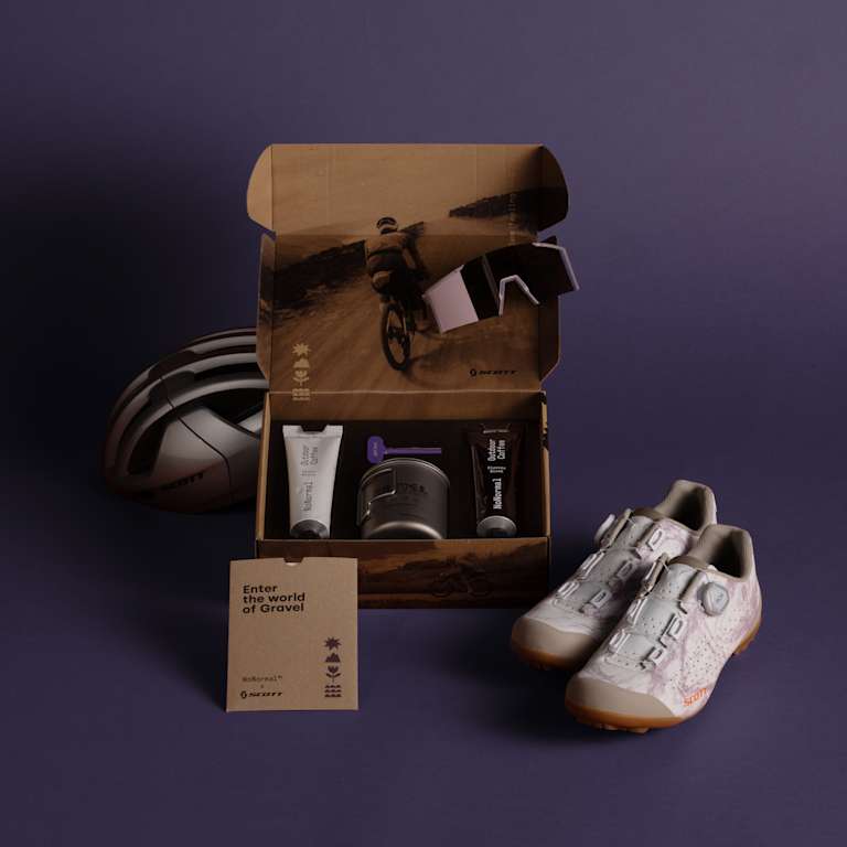 A cycling kit with shoes, helmet, and grooming products is arranged on a purple background, with an open box revealing more items and a scenic cycling image inside.