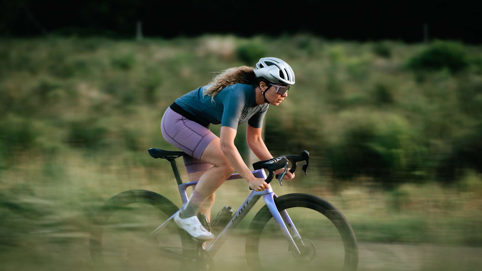 A cyclist rides on a dirt path through a sun-drenched field with trees in the background.