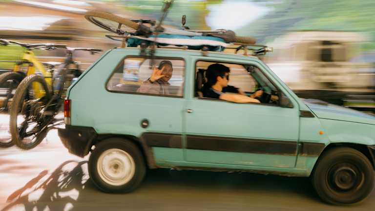 A light blue car with bicycles on its roof and back drives past a blurred background of trees and buildings.