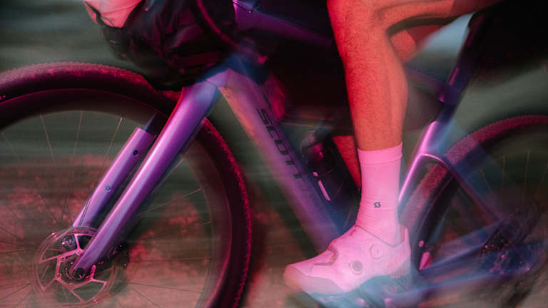 A cyclist in pink socks and white shoes pedals a purple bike with a blurred background of a dusty path.