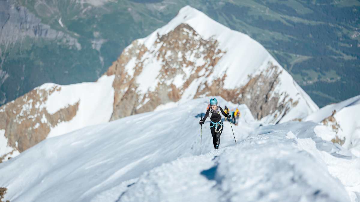 Iris Pessey ascending a steep snow ridge on Mont Blanc, trekking poles in hand with a sweeping alpine panorama behind her