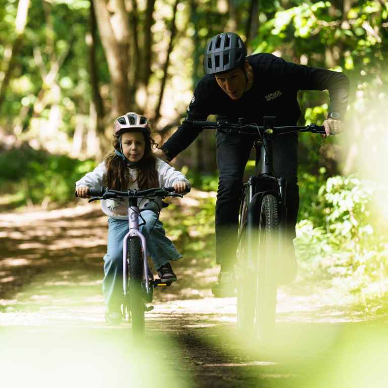 A child and adult ride a bike on a sunlit path surrounded by vibrant pink and purple flowers. The scene is joyful and filled with lush greenery.