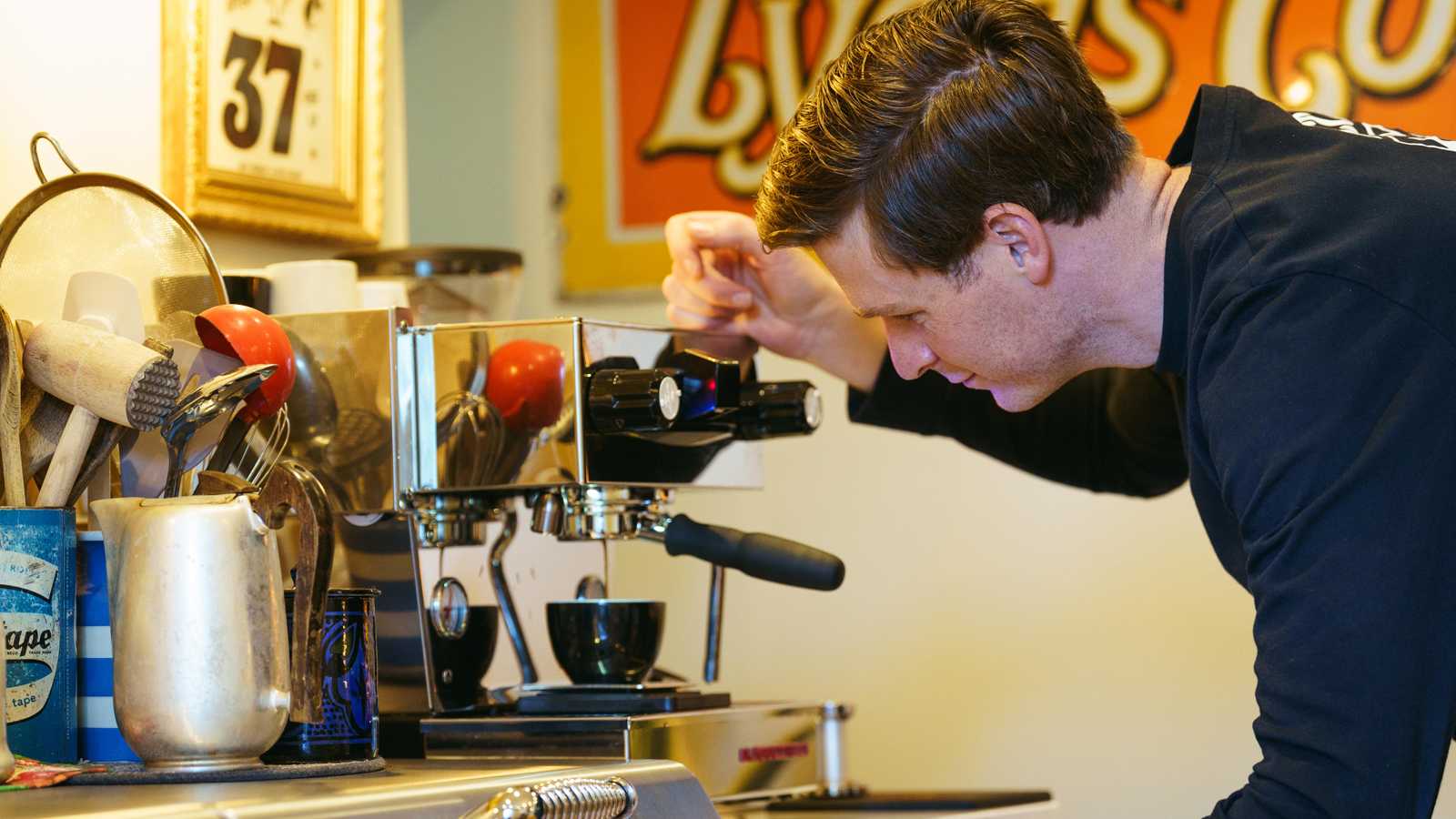 Man operating a shiny chrome espresso machine in a cafe with various kitchen tools visible.