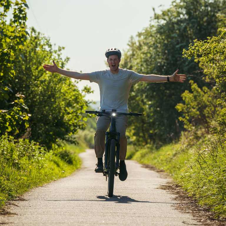 Joyful man with arms outstretched rides an e-bike down a sun-dappled path lined with lush green trees.