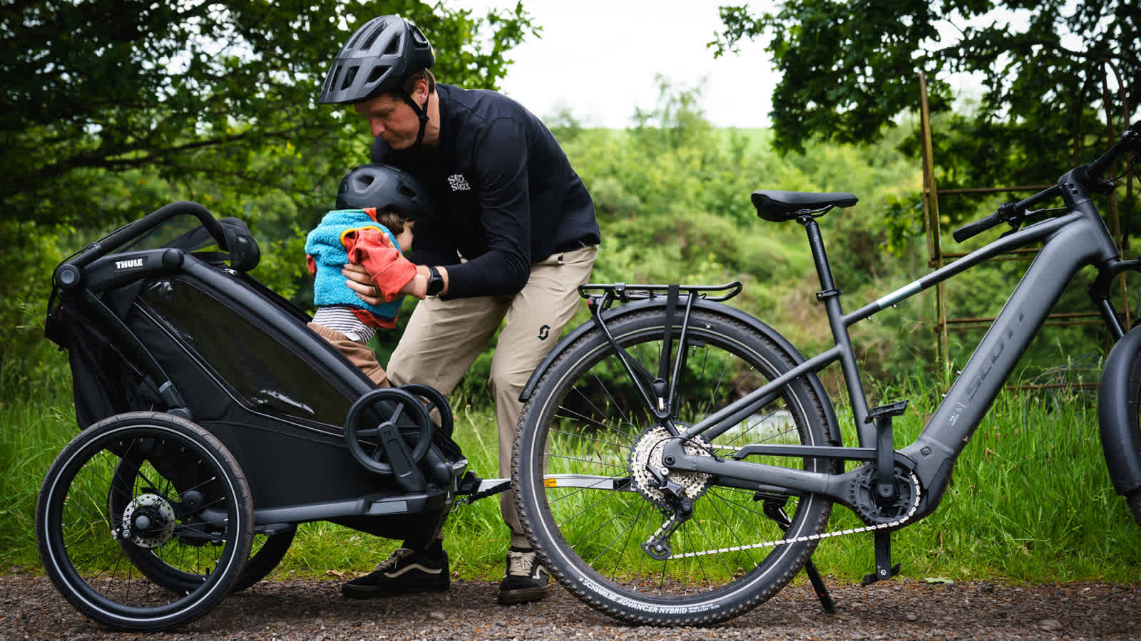 Man in helmet adjusts child in bike trailer next to a grey electric bicycle.
