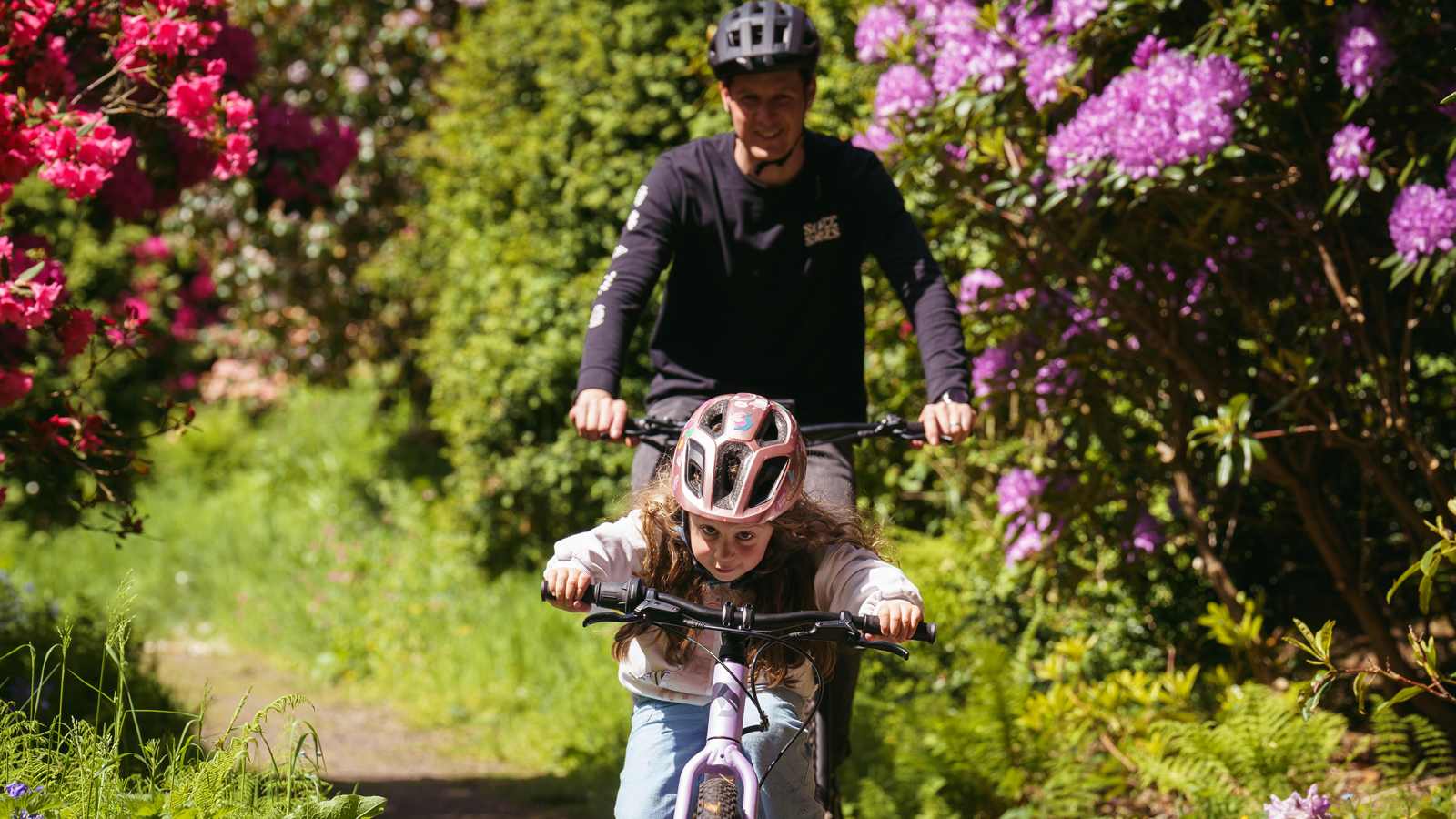 A child and adult ride a bike on a sunlit path surrounded by vibrant pink and purple flowers. The scene is joyful and filled with lush greenery.