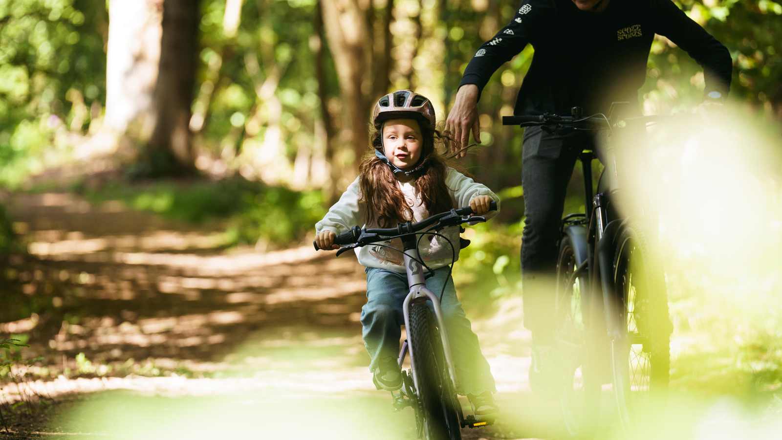 A young child wearing a helmet rides a bicycle on a forest path, accompanied by an adult on another bike in the background.
