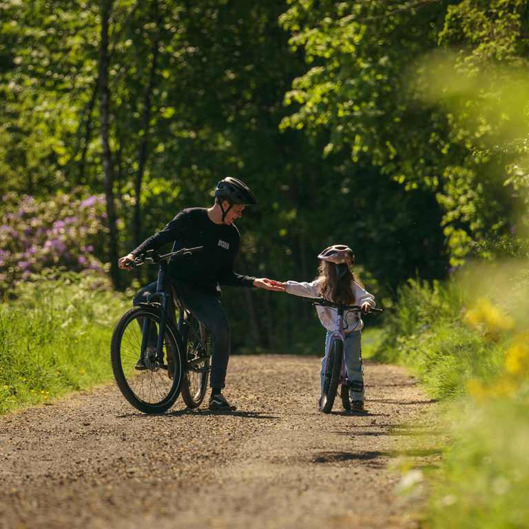 A man and a child riding bicycles together along a scenic path surrounded by greenery.
